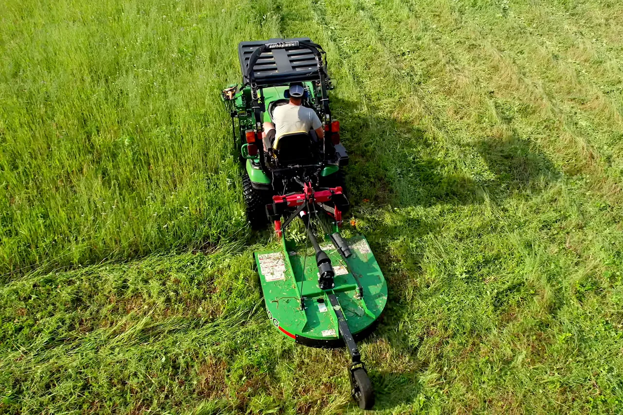A compact tractor pulling a rotary cutter through tall grass, seen from above